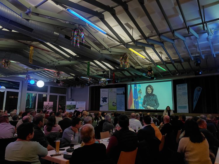 Großer Saal mit Publikum, das auf eine Leinwand mit einer Frau blickt, bunte Deckenbeleuchtung und hängende Dekorationen sichtbar.