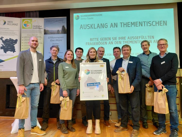Gruppe von neun Personen steht vor einer Leinwand mit Text und hält braune Papiertüten, eine Person hält ein Schild mit der Aufschrift 'SCHWAMMREGION Streu-Saale'.