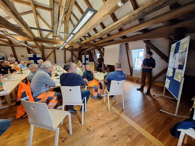 Mehrere Personen sitzen in einem Raum mit sichtbaren Holzbalken an der Decke und hören einem stehenden Mann zu, der vor einer Pinnwand spricht