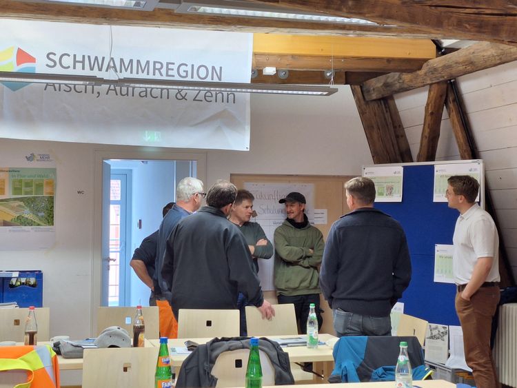 Interaktive Gruppenarbeit der Bauhöfe: Gruppe von sechs Männern in einem Raum mit Holzbalken, die vor Informationsplakaten und einem Banner mit der Aufschrift 'SCHWAMMREGION Aisch, Aurach & Zenn' stehen und sich unterhalten