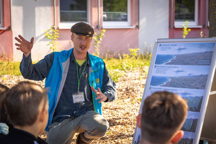 Mann mit blauer Weste und schwarzer Mütze erklärt zwei Kindern draußen vor einem Gebäude an einer Infotafel Vegetation
