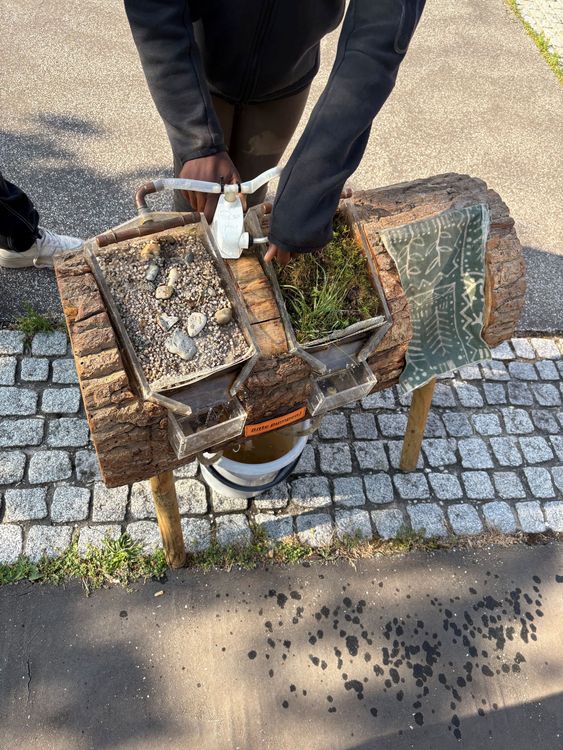 Holzblock mit zwei rechteckigen Pflanzkästen, einer mit Erde und Steinen, der andere mit Pflanzen, darüber eine Wasserhahnarmatur, Hände bedienen den Hahn:Versickerungsmodell am Aktionstag Wasser an der Mittelschule Emskirchen